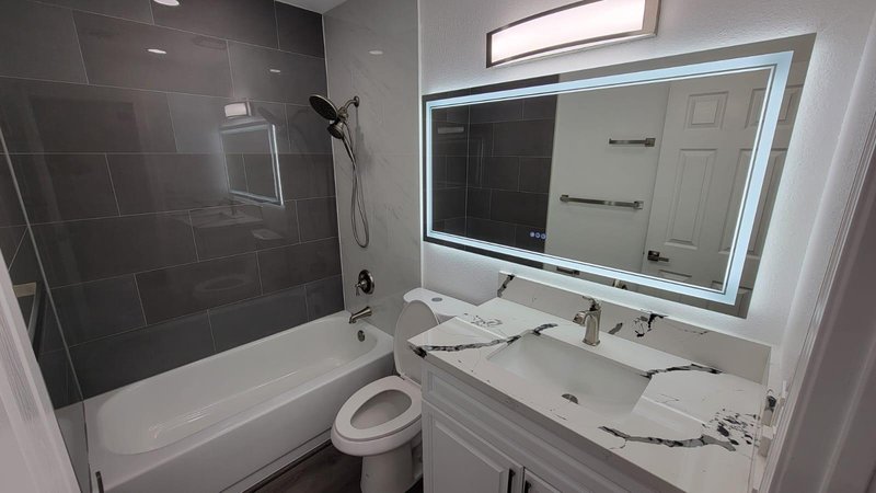 Baño con ducha de tile gris oscuro, tina y espejo LED sobre countertop de cuarzo